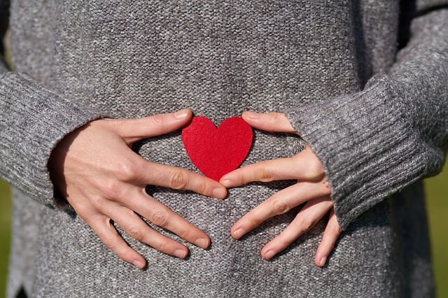 Frau mit grauem Pullover hält ein rotes Herz vor ihren Bauch